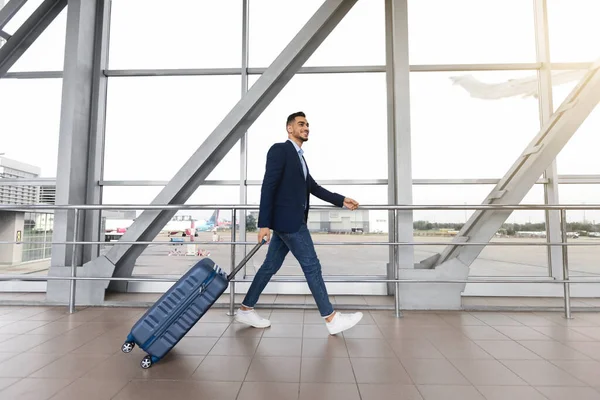 Retrato del hombre de negocios árabe sonriente con la maleta caminando cerca de la ventana en el aeropuerto — Foto de Stock