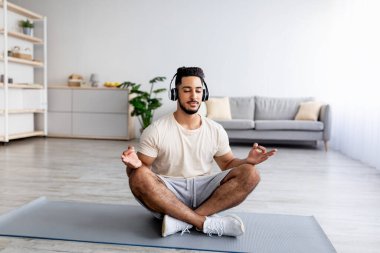 Nilüfer çiçekli poz veren, kulaklık takan, evdeki paspasın üzerinde müziği rahatlatmak için meditasyon yapan genç bir Arap.