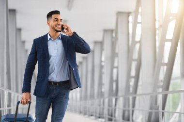 Orta Doğulu genç bir işadamı havaalanında çantayla yürüyor ve cep telefonuyla konuşuyor.