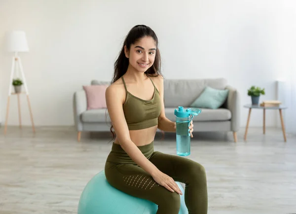 Atletisk ung indisk kvinna håller flaska vatten medan du sitter på fitness boll hemma — Stockfoto