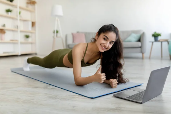 Sports en ligne. Femme indienne millénaire debout dans la planche de coude devant l'ordinateur portable, montrant pouce vers le haut geste à la maison — Photo