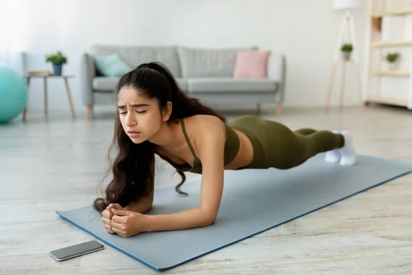 Utmattad ung indisk dam gör planka, tittar på mobiltelefon skärm, har svårt under styrketräning — Stockfoto