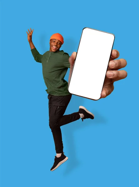 Fešák mladý černošky chlap držící smartphone, ukazující prázdné obrazovky, skákání přes modré studio pozadí, mockup — Stock fotografie