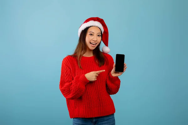 Christmas mobile offer. Happy asian lady in Santa hat pointing at smartphone with blank screen, blue background, mockup — Stock Photo, Image