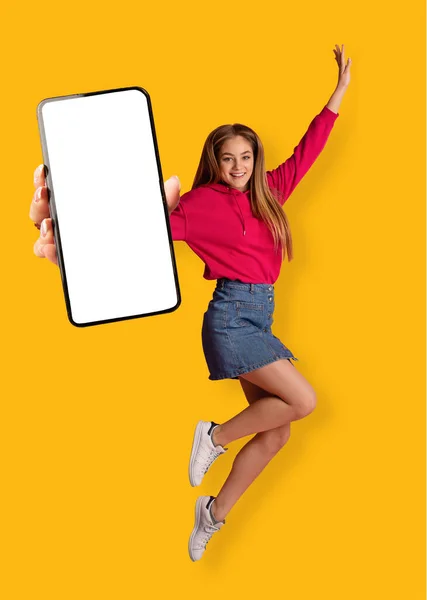 Mujer blanca alegre saltando en el aire, mostrando teléfono inteligente con pantalla blanca en blanco sobre fondo naranja, maqueta — Foto de Stock