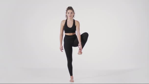 Jeune femme sportive souple portant costume de sport noir levant une jambe de côté, équilibrage sur fond de studio blanc — Video