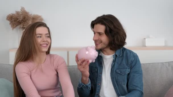 Közös családi megtakarítás. Fiatal boldog házastársak hallgatni teljes malac bank zaj, élvezi költségvetés együtt és nevetés — Stock videók