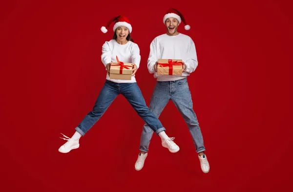 Emocionada pareja milenaria que usa sombreros de Papá Noel saltando con regalos de Navidad en las manos — Foto de Stock