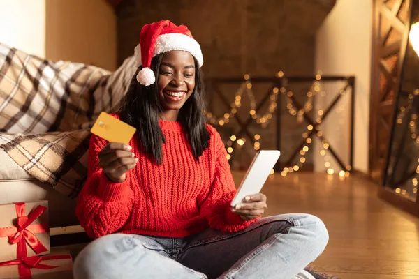 Felice signora nera con carta di credito utilizzando la nuova app di shopping sul telefono cellulare per acquistare regali di Natale da casa, copia lo spazio — Foto Stock