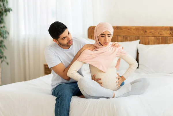 Těhotná muslimka žena má bolesti při porodu, sedí po boku manžela na posteli, arabky muž masíruje manželky — Stock fotografie
