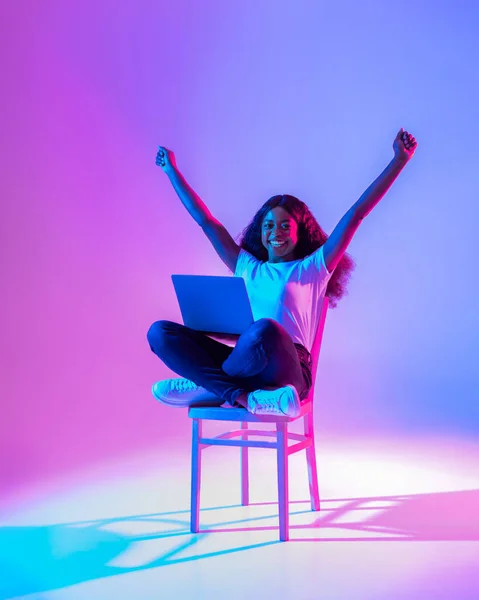 Emocionado joven dama negra sentada en la silla con el ordenador portátil, levantando los brazos, celebrando el éxito en la luz de neón — Foto de Stock