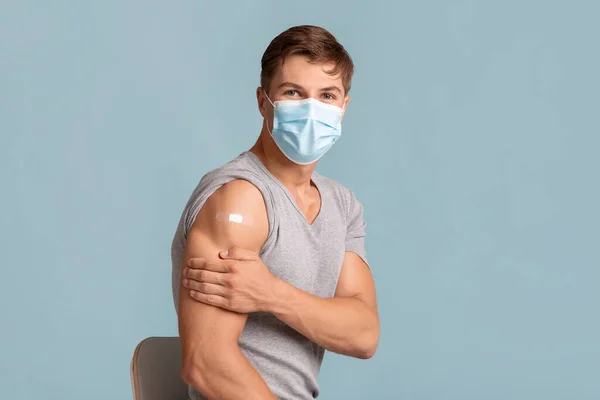 Šťastný mladý běloch v ochranné masce show rameno s náplastí po injekci izolované na modrém pozadí — Stock fotografie