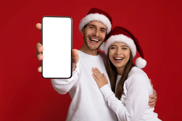 Super appli. Heureux jeune couple portant des chapeaux de Père Noël montrant écran Smartphone vide — Photo