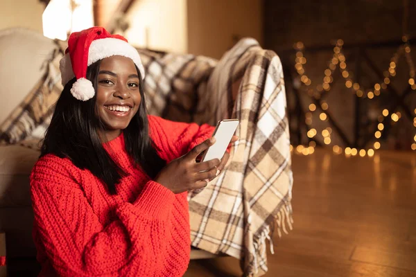 Felice signora nera in maglione di Natale e cappello di Babbo Natale utilizzando il telefono cellulare, shopping online, la selezione di regali a casa, spazio copia — Foto Stock
