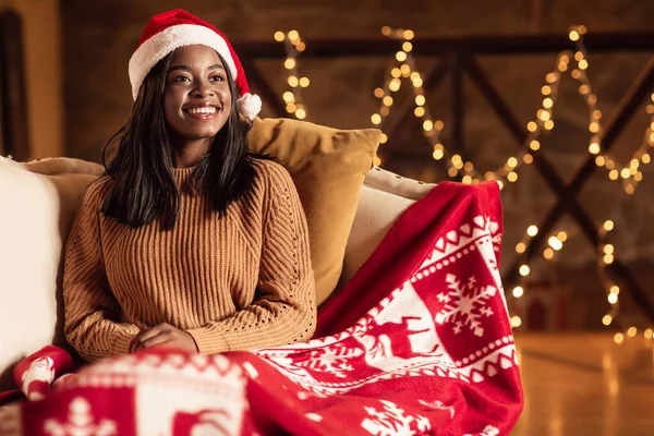 Accogliente atmosfera natalizia. Giovane donna nera in cappello Babbo Natale seduta sul divano sotto il caldo plaid, che celebra Natale a casa — Foto Stock