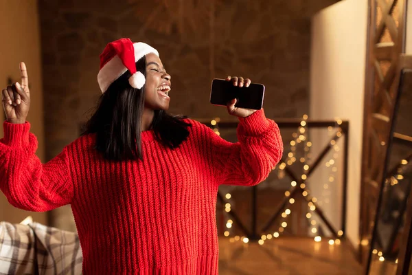 Musica natalizia. Felice signora nera in Santa cappello canto, utilizzando smartphone come microfono a casa, mockup per l'applicazione mobile — Foto Stock