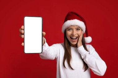 Harika bir teklif. Noel Baba Şapkalı Heyecanlı Genç Bayan Boş Akıllı Telefon Gösteriyor