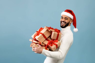 Kutlama zamanı. Noel Baba şapkalı neşeli Arap adam elinde hediye kutuları, mavi arka planda poz veriyor.