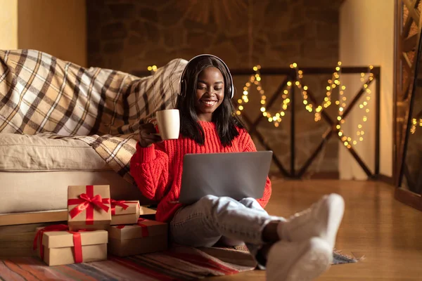 Black woman celebrating Christmas alone, using laptop, watching Xmas movie, drinking coffee near gift boxes at home — Stock Photo, Image