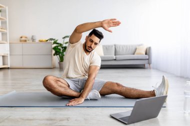 Tam boy atletik genç Arap adam oturma odasında bacağını yoga minderine uzatıyor.