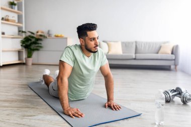 Yurt içi yoga pratiği. Esnek genç Arap adam minder üzerinde kobra pozu veriyor, evde egzersiz yapıyor, boş yer var.