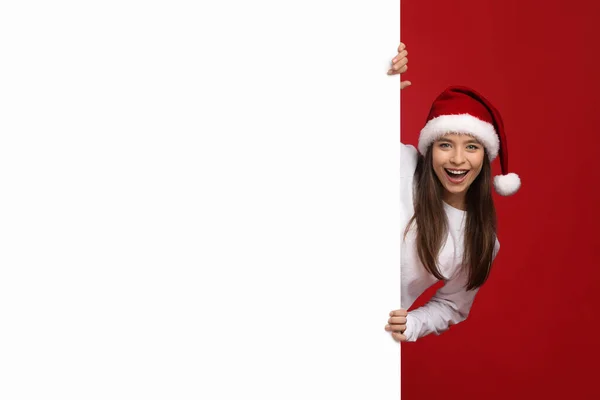 Vánoční prodej. Nadšený dáma v Santa Klobouk nakukování za bílou reklamní tabuli — Stock fotografie