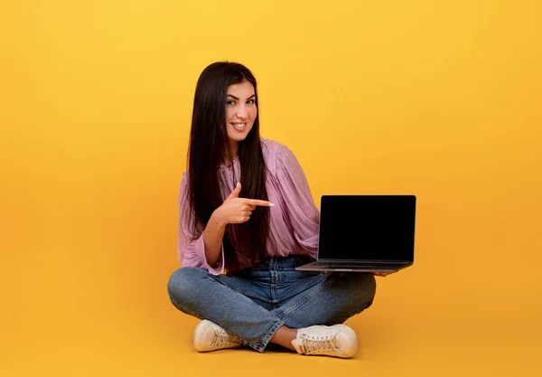 Sitio web o anuncio fresco. Mujer armenia feliz apuntando a la computadora portátil con pantalla en blanco, sentado en el fondo amarillo — Foto de Stock
