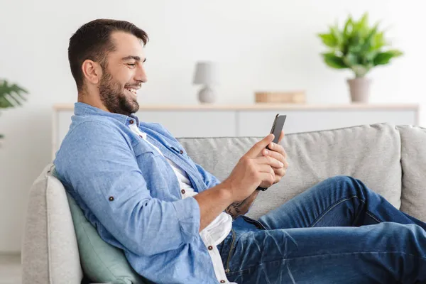 Alegre adulto europeu cara com barba senta-se no sofá conversando no telefone na sala de estar interior, vista lateral — Fotografia de Stock