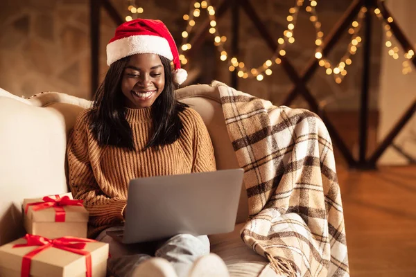 Donna nera con computer portatile seduto sul divano che saluta le e-mail di Natale online in atmosfera di casa vacanza — Foto Stock