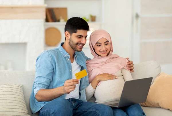 Prepararsi per il parto. Amare musulmano coppia incinta utilizzando computer portatile e carta di credito, seduto sul divano — Foto Stock