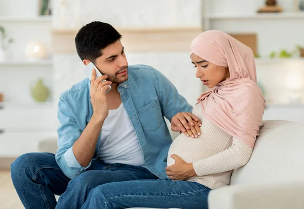 Těhotná arabská žena s prenatální kontrakce, ustaraný manžel volá lékaře, pomocí mobilního telefonu, domácí interiér — Stock fotografie