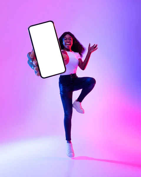 Lunghezza completa della donna nera eccitata che salta con il cellulare, mostrando uno schermo vuoto nella luce al neon, mockup per sito Web o app — Foto Stock