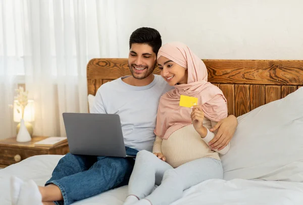 Giovani musulmani in attesa di famiglia con computer portatile e carta di credito, shopping online mentre seduti sul divano a casa — Foto Stock