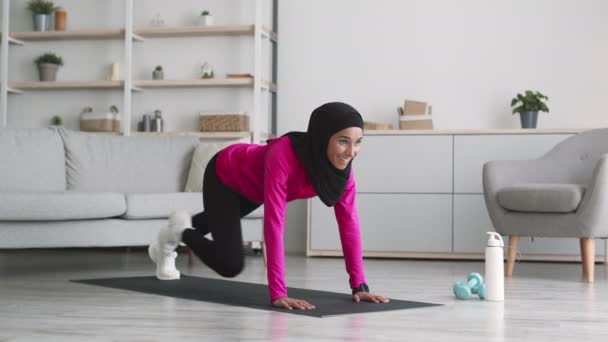 Joven mujer musulmana activa practicando la tabla de correr en la alfombra de fitness en casa, disfrutando del entrenamiento de escalador de montaña y sonriendo — Vídeo de stock