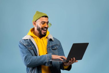 Serbest meslek hayatı. Heyecanlı Arap adam dizüstü bilgisayar kullanıyor ve mavi arka planda dururken gülümsüyor.