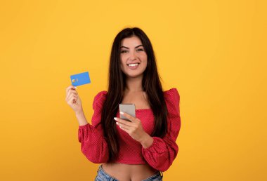 Kolay para transferi. Neşeli Ermeni kadın kredi kartı gösteriyor ve elinde akıllı telefonu, sarı arka planı var.