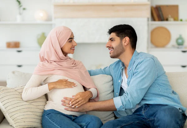 Glückliches muslimisches Paar erwartet Baby, ruht zu Hause auf Couch und lächelt, Mann berührt Schwangeren den Bauch — Stockfoto