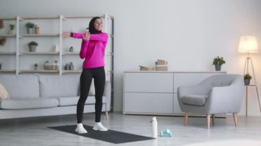 Genç İslami Ortadoğulu tesettürlü bayan evde sabah keyfine bakıyor. Fiziksel serseriler, ısınıyor ve kollarını uzatıyor.