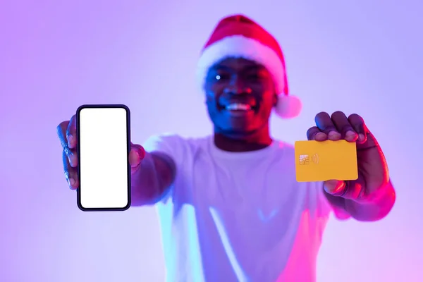 Joyeux jeune homme noir à Noël Santa chapeau montrant carte de crédit et smartphone avec écran blanc dans la lumière au néon, maquette — Photo