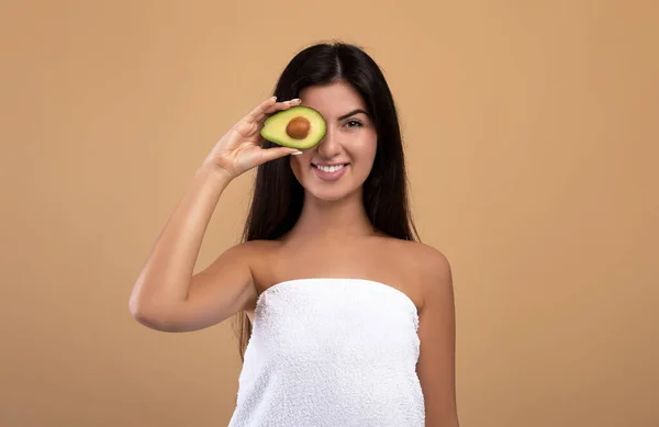 Concepto de cuidado de la piel orgánico. Feliz dama armenia sosteniendo la mitad de aguacate fresco y cubriendo el ojo, posando sobre fondo beige — Foto de Stock