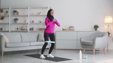 Yoğun aile eğitimi. Genç ortadoğulu bayan evde antrenman yapıyor, elastik bantla bacaklarını kaldırıyor.