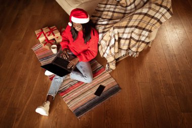 Noel Baba şapkalı mutlu siyah kadın evde web sitesi için dizüstü bilgisayarı kullanıyor, Xmas hediyeleri alıyor, üstten bakıyor.