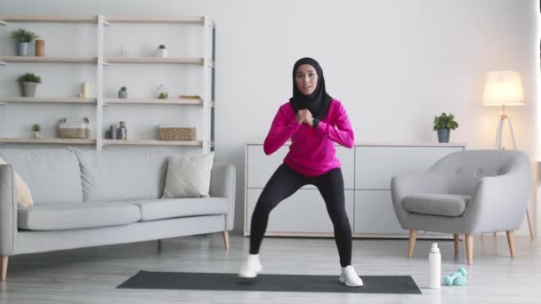 Joven mujer activa de Oriente Medio en el entrenamiento de hijab en casa, haciendo sentadillas activas en casa y sonriendo, cámara lenta — Vídeo de stock