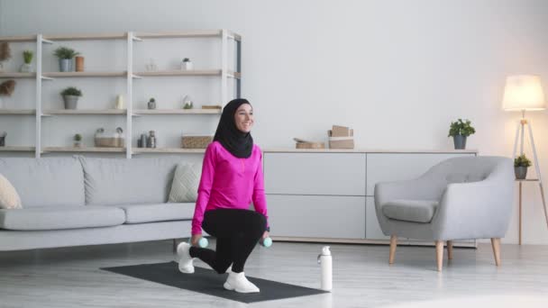 Joven mujer activa de Oriente Medio entrenando en casa, practicando ataques hacia adelante ejercicio con mancuernas y sonriendo — Vídeo de stock