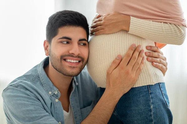 Junger arabischer Mann legt sein Ohr auf muslimischen Ehefrauen schwangeren Bauch, hört Babys Herzschlag, Nahaufnahme — Stockfoto