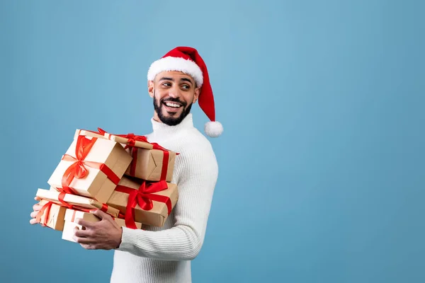 Días festivos y tiempo de celebración. Chico árabe feliz en Santa sombrero sosteniendo regalos y mirando el espacio libre, fondo azul — Foto de Stock
