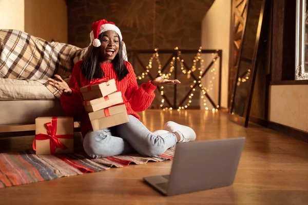 サンタの帽子を身に着けているクリスマスの贈り物と興奮した黒人女性,ラップトップ上でビデオチャットを持っています,クリスマスとお祝いの友人 — ストック写真