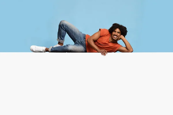 Happy black guy relaxing on huge empty placard over blue — Stock Photo, Image