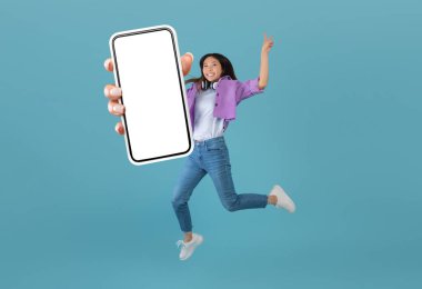 Heyecanlı Asyalı kadın mavi stüdyo arkaplanı, kolaj, afiş üzerinden atlarken boş akıllı telefon ekranı gösteriyor.
