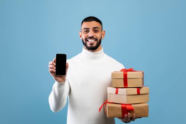 Mutlu Arap adam cep telefonunu boş ekran ile gösteriyor ve elinde hediyeler var, mavi stüdyo arka planında duruyor.